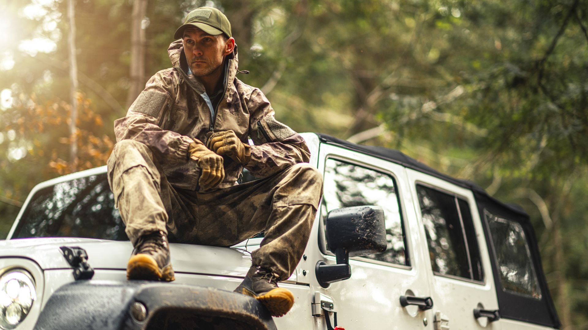 Soldier in camouflage sits on white Jeep in forest with sunlight